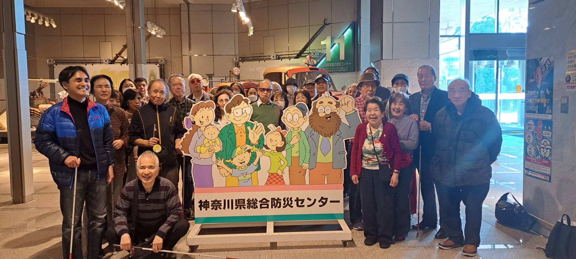 多くの人々が集まって記念撮影をしています。彼らの前には「神奈川県総合防災センター」と書かれた看板があります。看板には、家族のイラストが描かれています。背景には展示物が見え、建物の中で撮影されたようです。参加者の中には、白杖を持っている人もいます。皆さんが笑顔で楽しそうにしています。