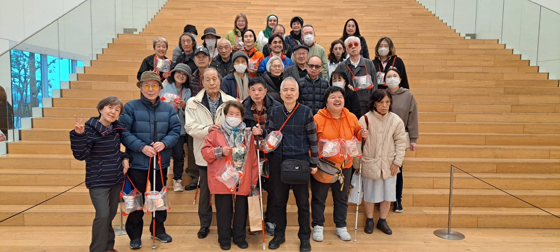 階段の前で集団写真を撮っている人々のグループ。年配の方々が多く、皆さん笑顔でカメラを見ています。多くの人が白杖を持っており、首から赤いストラップのついた透明なバッグを下げています。