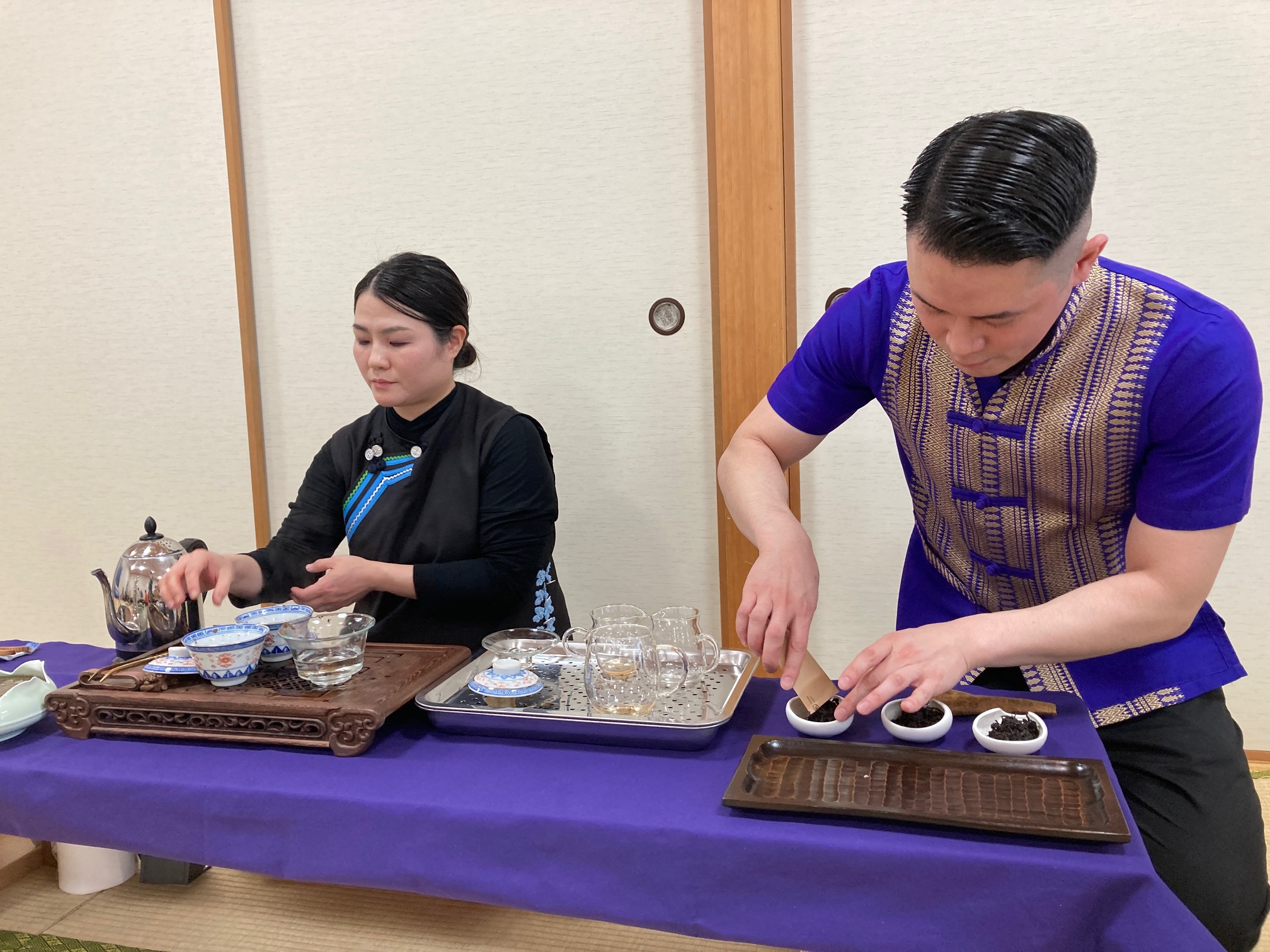 2人の人物がテーブルでお茶を準備しています。左側の女性は黒い服を着ており、ティーポットを持っています。右側の男性は青い服を着ており、小さなボウルに茶葉を入れています。テーブルには紫色の布が敷かれており、いくつかのティーカップとガラスの容器が並べられています。